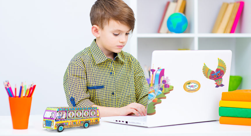 A boy sitting at his desk working at his computer which has Crayola imagine arts academy decals on the back. on the desk is a