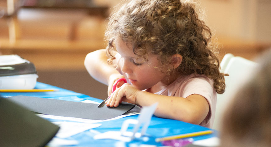A girl sitting at a desk using scissors to cut a black piece of black construction paper