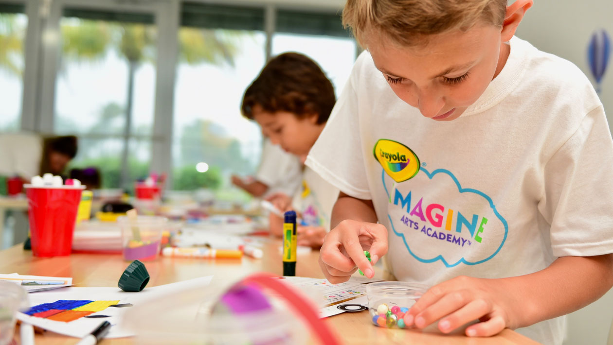 Blonde boy wearing a Crayola imagine arts academy tshirt coloring in an art class